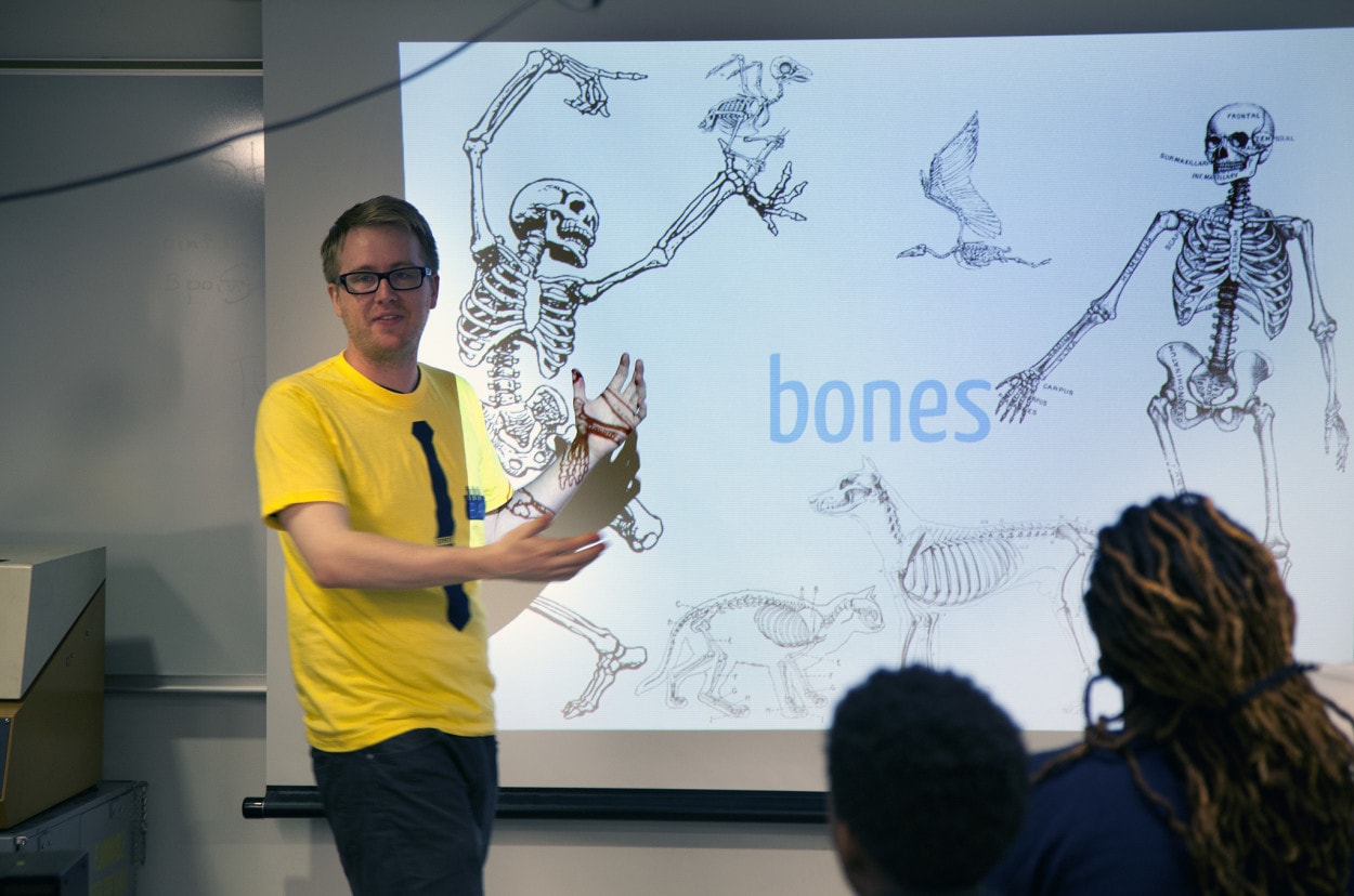 Blond man in a maize shirt stands in front of a smartboard with bones displayed on it