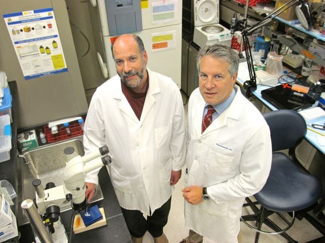 Two white males stand in white lab coats in a lab
